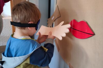 Enfant avec les yeux bandés qui tient une main en carton devant un visage en carton dessiné  - Agrandir l'image 1 sur 3, fenêtre modale