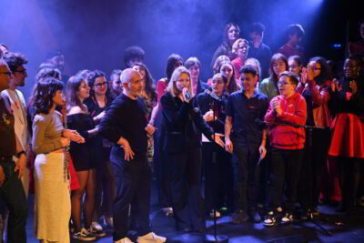 Jeune fille qui chante avec un micro entourée d'autres personnes sur une scène - Agrandir l'image 2 sur 3, fenêtre modale
