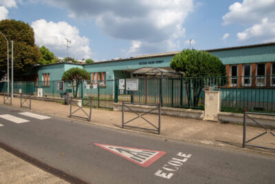 Bâtiment bleu claire avec des fenêtre en bois et un grand portail vert. Il s'agit de l'école Jules Ferry.  - Agrandir l'image 5 sur 9, fenêtre modale